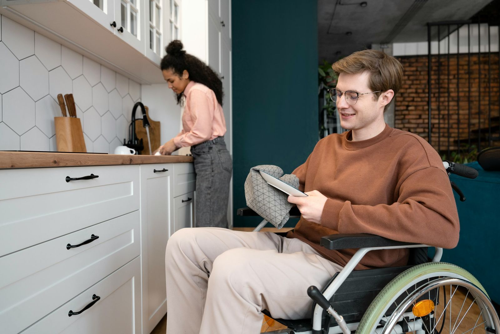 Un jeune homme en fauteuil roulant feuillette un document dans sa cuisine pendant qu'une femme s'affaire au plan de travail.