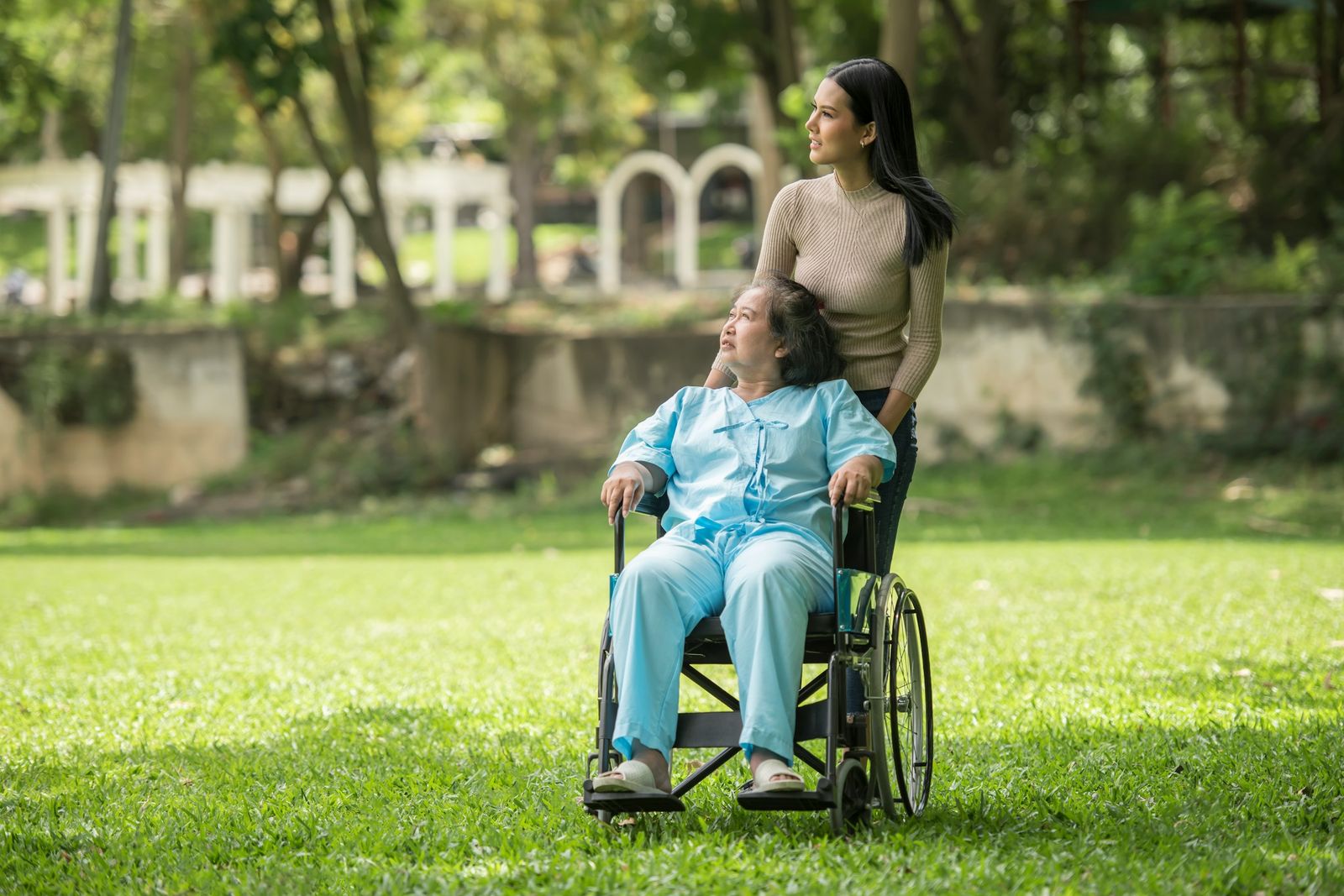 Dans un parc, une jeune femme pousse le fauteuil roulant d'une femme plus âgée, qui lève les yeux vers elle.