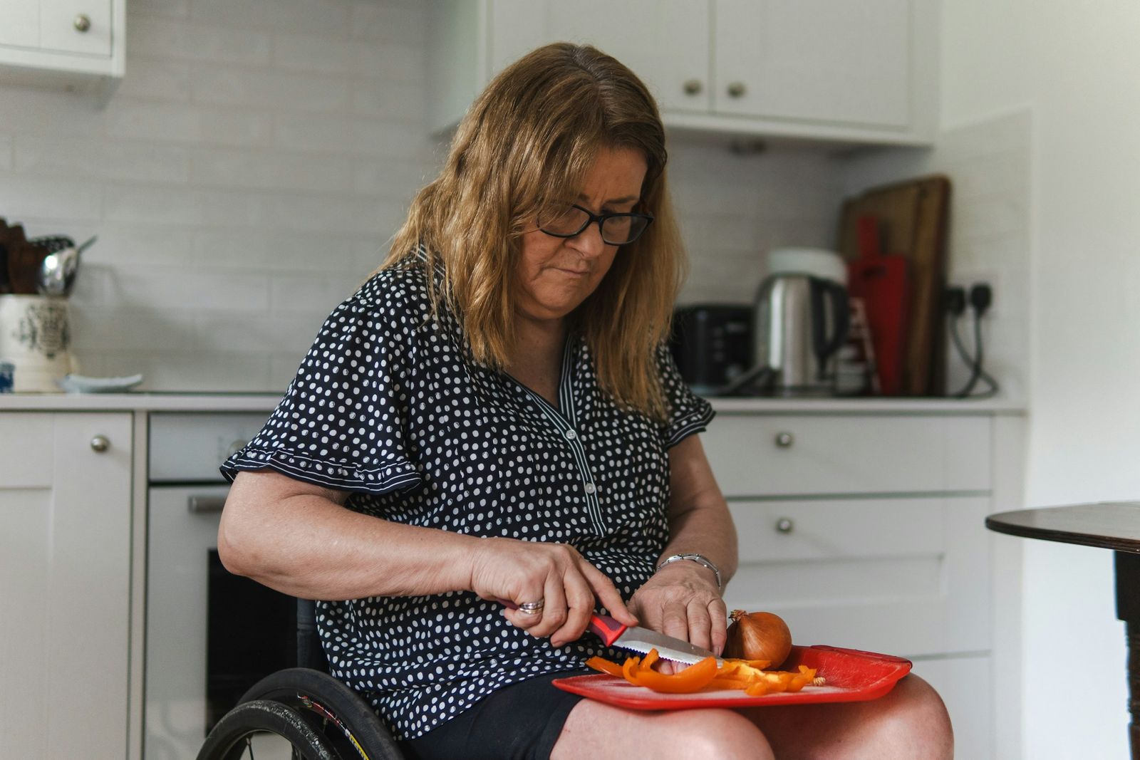 Dans sa cuisine, une femme en fauteuil roulant coupe des légumes sur un plateau posé sur ses genoux.