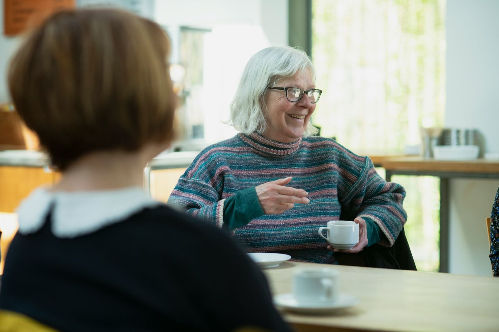 Une femme âgée, une tasse à la main, discute en souriant avec une autre femme vue de dos.