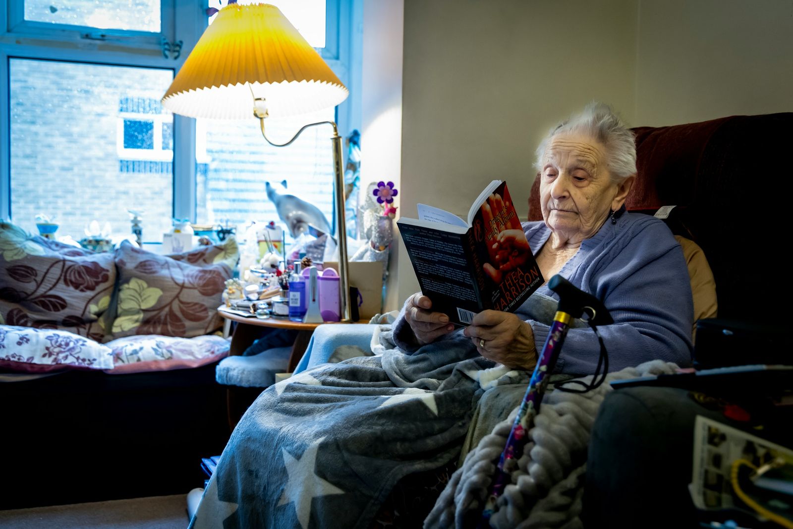 Une femme âgée lit dans son fauteuil, une couverture sur les genoux et sa canne à portée de main.