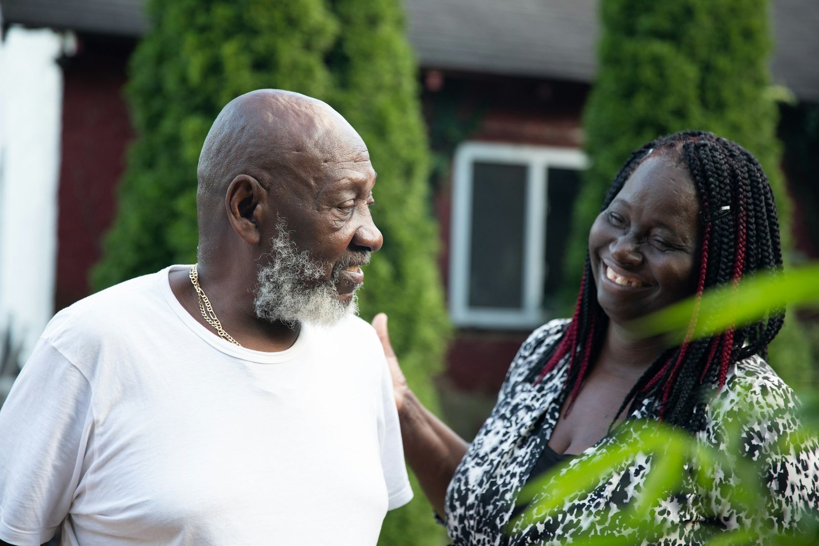 Devant une maison, une femme pose la main sur l'épaule de son père âgé ; ils échangent un sourire.